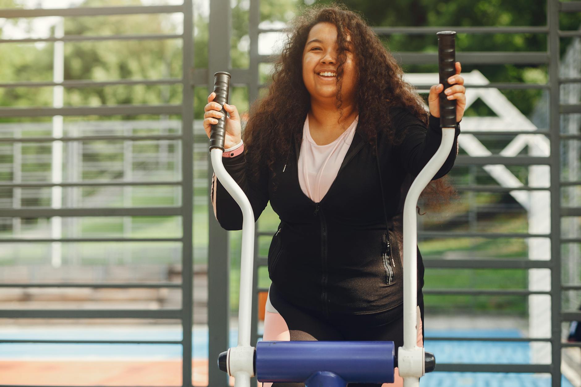 Happy woman with curly hair enjoying exercise on an outdoor elliptical machine in the park.
